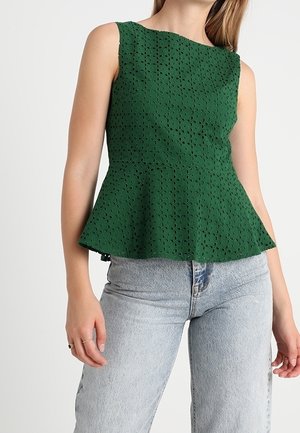 Zielony top peplum wykonany z fakturowanej tkaniny z wyciętymi wzorami, z dekoltem typu łódka i bez rękawów, zestawiony z jasnoniebieskimi dżinsami.