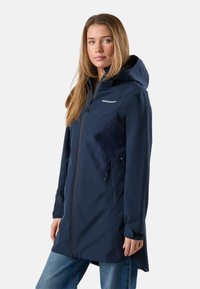 Kvinde iført en marineblå hættetrøje med lynlås foran og sidelommer, kombineret med blå jeans, stående mod en ensfarvet baggrund.