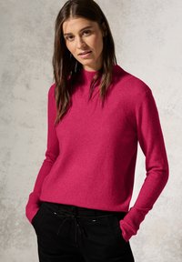 Enganliegender, gerippter Rollkragenpullover in leuchtendem Pink, aus weichem Strickstoff, kombiniert mit schwarzen Jogginghosen mit Kordelzug.