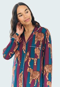 Camicia da pigiama a maniche lunghe con stampa di tigre arancione su uno sfondo a righe di un bordeaux scuro e teal, dotata di taschino e colletto con pizzo.