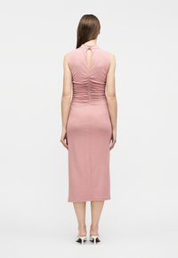 Γυναίκα  που στέκεται με την πλάτη γυρισμένη, φορώντας ροζ μίντι φόρεμα χωρίς μανίκια με ρυτιδιασμένη πλάτη και λαιμόκοψη με άνοιγμα, λευκά ψηλοτάκουνα mules σε απλό φόντο.