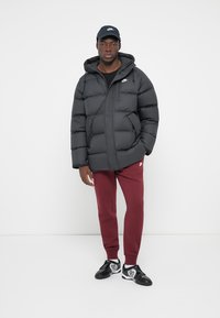 Sort pufferjakke med lynlås foran, hætte og sidelommer, parret med bordeauxfarvede sweatpants og sorte sneakers med hvide accenter.