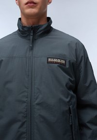 Giacca zip-up grigia scura con colletto alto, dettagli cuciti e una toppa con marchio sul petto. Tessuto liscio e leggero.