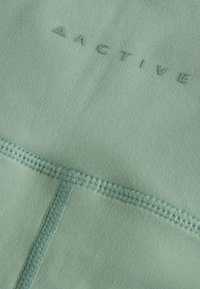 Sporttop in muntgroen, met een gladde textuur, flatlocknaden en een reliëflogo.