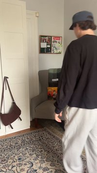 Personne en chemise noire et pantalon gris marchant près d'un fauteuil gris avec une couverture à carreaux et un livre coloré, près d'une porte avec un sac marron suspendu.