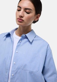 Chemise bleu clair à manches longues avec des rayures verticales blanches et rouges, col évasé, poche poitrine unique et texture en coton lisse.