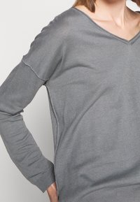 Maglione grigio a maniche lunghe con scollo a V, texture morbida e spacchi laterali. Presenta spalle cadute e polsini a coste, evidenziando una vestibilità rilassata.