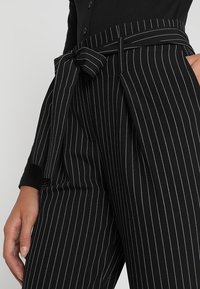 Black high-waist pinstripe pants with white vertical lines and a fabric belt tied in a bow at the waist, paired with a black buttoned top.