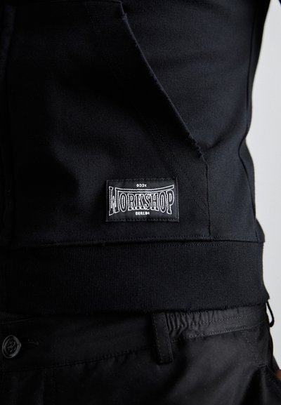 Black sweatshirt with a ribbed hem and a sewn label reading "WORKSHOP BERLIN." Features a soft fabric texture and a fitted design.