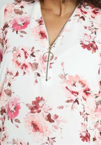 Blusa floreale bianca, con motivi floreali rosa e marroni, scollo a V e cerniera dorata con tiretto circolare.