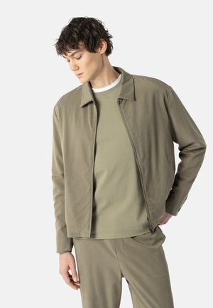Ung mand iført koordineret let olivengrøn jakke, matchende bukser og en lysere olivengrøn skjorte, poser med hænderne tæt på lommerne mod en hvid baggrund.