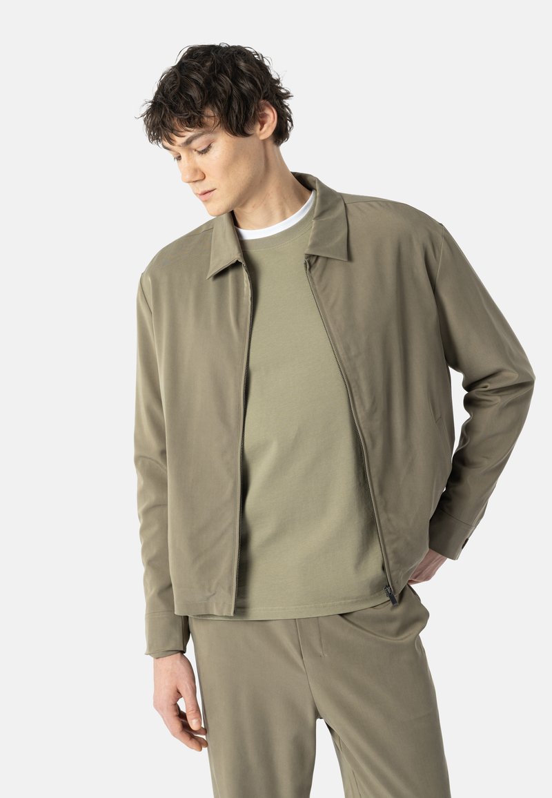Jeune homme portant une veste kaki clair assortie, un pantalon assorti et une chemise kaki plus claire, posant avec les mains près des poches sur un fond blanc.