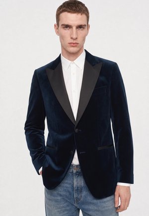 Junger Mann trägt einen dunkel samtigen Blazer mit schwarzen Revers, weißem Hemd und blauen Jeans, steht mit einer Hand in der Tasche.