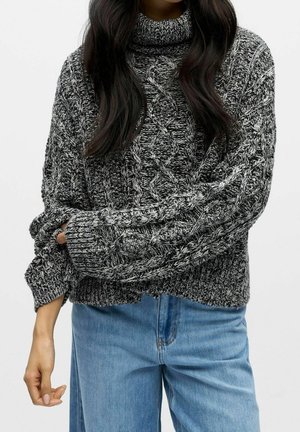 Mujer con un suéter de cuello alto de punto con cable en blanco y negro y jeans azul claro, con el cabello largo y oscuro, de pie frente a un fondo liso.