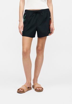 Femeie purtând pantaloni scurți negri cu talie elastică cu broderie florală subtilă, top alb și sandale maro țesute, stând pe un fundal alb.