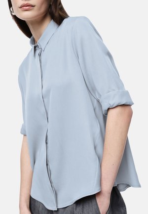 Donna con camicia azzurra a maniche lunghe abbottonata, maniche arrotolate, in piedi con una mano in tasca contro uno sfondo semplice.