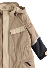 Tan buitenjack met een beige voorstreep, een grote zak, zwarte mouwaccenten en een kap gevoerd met zachte stof. Inclusief reflecterende details.