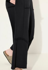Pantaloni neri a gamba larga realizzati in un tessuto morbido, con cuciture sfilacciate, grandi tasche e una vestibilità rilassata. Abbinati a scarpe slip-on di colore chiaro.