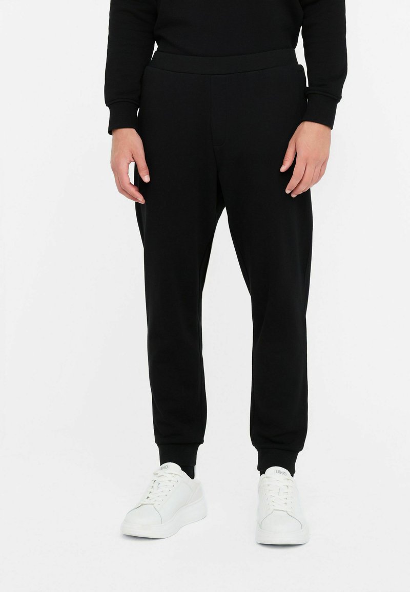 Sorte joggingbukser med elastisk liv, tapered ben og sidelommer, parret med hvide sneakers. Blød stofftekstur.