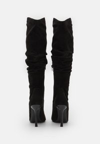 Bottes montantes en daim noir avec une coupe ajustée, un léger froncement subtil et des talons pointus. Dotées d'une fermeture éclair latérale pour faciliter l'enfilage.