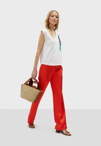 Top bianco senza maniche caratterizzato da ricami colorati, abbinato a pantaloni in satin larghi e rossi. La modella tiene una borsa di paglia intrecciata e indossa scarpe aperte.