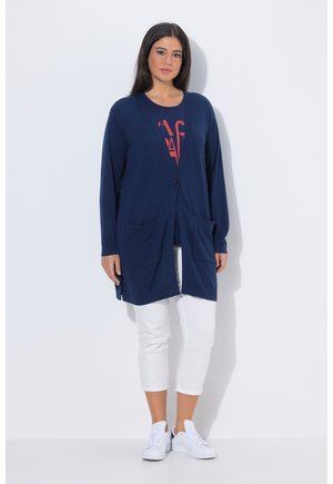 Donna che indossa un cardigan lungo blu navy sopra una maglietta rossa con grafica, pantaloni bianchi corti e scarpe da ginnastica bianche, in piedi contro uno sfondo bianco.