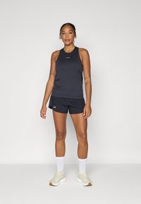 Top sport negru cu design tip racerback și logo, echipat cu pantaloni scurți negri. Pantofi sport bej și șosete albe completează ținuta.