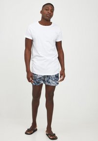Vit bomullst-shirt och marinblå badshorts med vita tropiska lövmönster. Svarta flipflops. Neutral bakgrund.