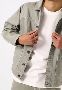 Giacca di jeans verde chiaro con chiusura a bottone, tasca sul petto e vestibilità rilassata, indossata sopra un maglione bianco. Trama morbida, design casual.