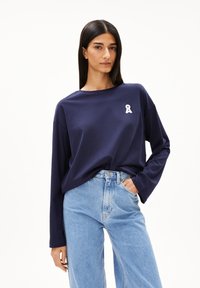 Marineblaues Langarmshirt aus weichem Stoff, mit einem kleinen weißen Ribbon-Logo auf der Brust, kombiniert mit hellblauen High-Waist-Jeans.