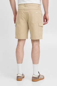 Beige cargo shorts laget av bomull med flere sidelommer, sydd sømmer og en avslappet passform, kombinert med lyse grå og hvite joggesko.