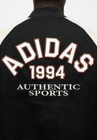 Μαύρο varsity μπουφάν με εκρού μανίκια που φέρει κεντημένο στο πίσω μέρος το κείμενο "ADIDAS 1994 AUTHENTIC SPORTS" με λευκά και πορτοκαλί γράμματα με περίγραμμα.