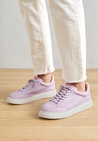 Sneakers di pelle viola con suola bianca, punta tonda e lacci piatti. Accentati da un logo rosso del marchio sulla linguetta. Jeans bianchi sfilacciati sullo sfondo.