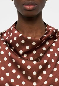 Gros plan du cou et des lèvres d'une personne portant un foulard en soie marron à pois blancs drapé autour des épaules.