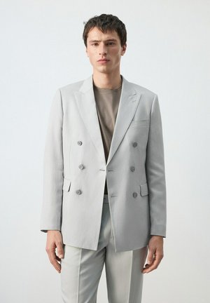 Młody mężczyzna ubrany w jasnoszary, dwurzędowy żakiet i spodnie oraz taupe koszulkę z okrągłym dekoltem, stojący na tle jednolitego, jasnego tła.