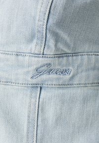 Vaalean sininen farkkukangas, jossa näkyvät ompeludetaljit. Kappaleessa lukee "Guess" kauniilla kursiivilla sauman varrella. Pintarakenteeltaan tekstuuri.