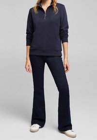 Marinblå zip-up sweatshirt med en texturerad bröstpanel, i kombination med mörka utsvängda jeans och ljusa sneakers mot en grå bakgrund.