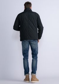 Giacca nera trapuntata con colletto alto, dettagli con bottoni e maniche lunghe, abbinata a jeans blu chiaro e stivali in camoscio beige.