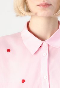 Ανοιχτό ροζ πουκάμισο με κλασικό γιακά και κόκκινες κεντημένες καρδιές. Κουμπί μπροστά με λεία υφή υφάσματος.