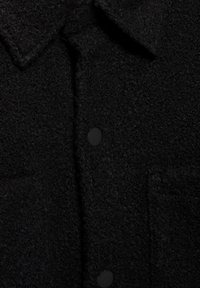 Sort tekstureret skjorte med en uldlignende overflade, med krave og to forlommer, lukket med sorte knapper.