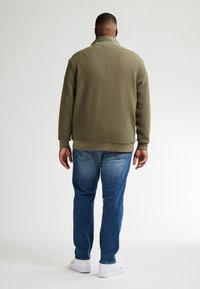 Olivgrön texturerad jacka med hög krage, i kombination med blå denimjeans och vita sneakers, sedd bakifrån.