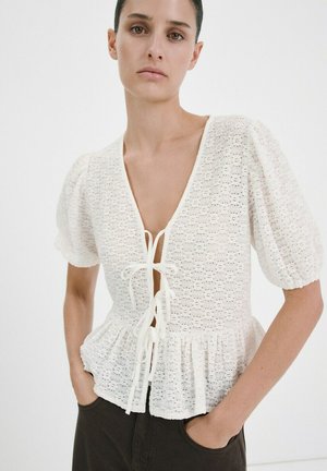 Žena oblečená v bielej textúrovanej blúzke s nafúknutými rukávmi a tromi prednými uzávermi na zaväzovanie, v kombinácii s tmavými nohavicami, na jednoduchom pozadí.