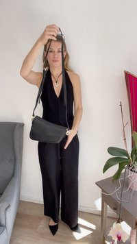 Black shoulder bag featuring a smooth texture, rectangular shape, and a long strap, held by a person in a black outfit against a neutral wall.