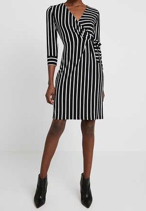 Kvinde iført en knælængde sort-hvidt, lodret striped slå-om kjole med 3/4 ærmer og sorte ankelstøvler.