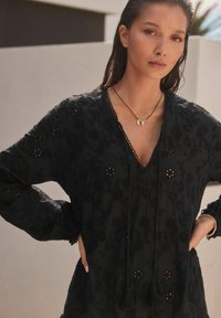 Tunique noire brodée avec des motifs floraux, décolleté en V, cordon de serrage avec des franges, manches longues, texture légère et détails élaborés.