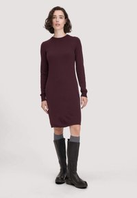 Burgunderfarbendes Strickkleid mit langen Ärmeln, Rundhalsausschnitt und taillierter Silhouette, kombiniert mit hohen schwarzen Stiefeln und grauen Socken.