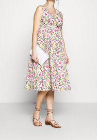 Vestido de estampado floral con escote en V, que presenta patrones en rosa, amarillo y verde. Combinado con sandalias marrones de tiras y un bolso de mano blanco.