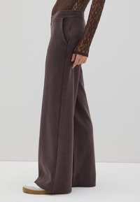 Pantalones de pierna ancha en tejido taupe oscuro con una textura suave, que presentan una cintura alta, bolsillos laterales y se combinan con zapatillas blancas y marrón claro.