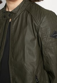 Olivgrön vadderad bomberjacka med ståkrage, dragkedja och svart logotypmärke på ärmen. Slätt, lätt material.