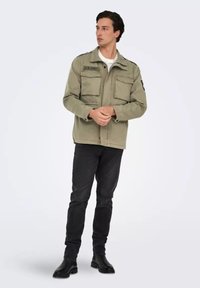 Giacca militare verde oliva con due tasche anteriori, chiusura con zip e toppe sulle spalle. Abbinata a jeans neri e stivali.